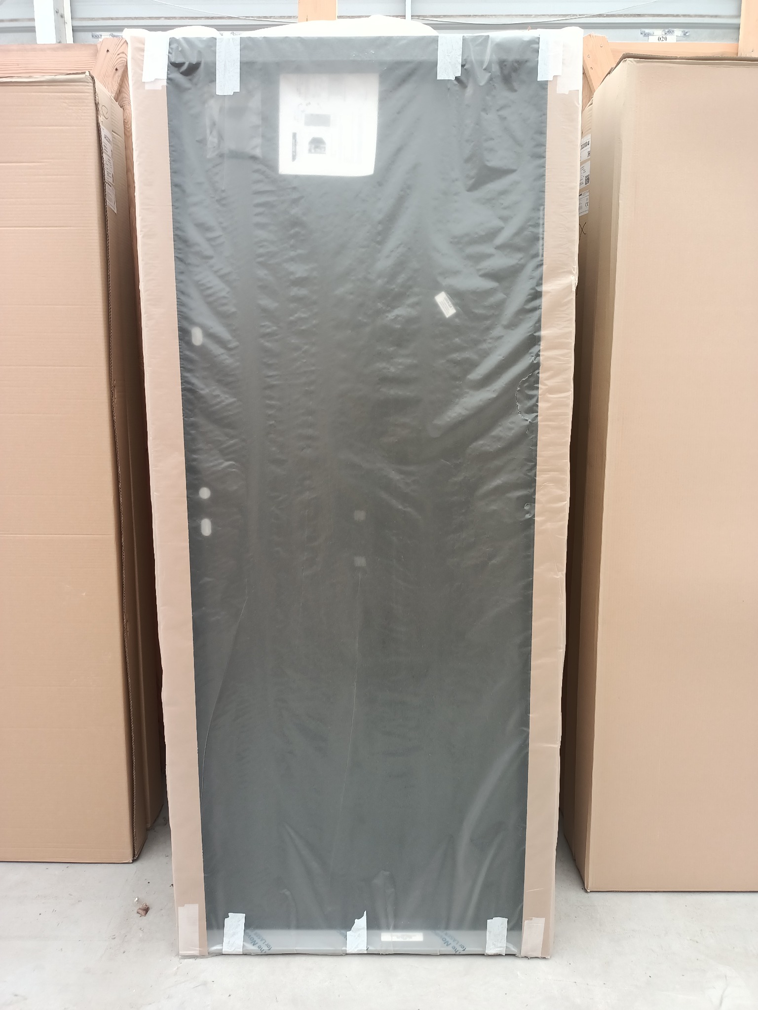 Nowe, zapakowane drzwi wewnętrzne w folii ochronnej i kartonowym zabezpieczeniu, ustawione pionowo między kartonami w magazynie.