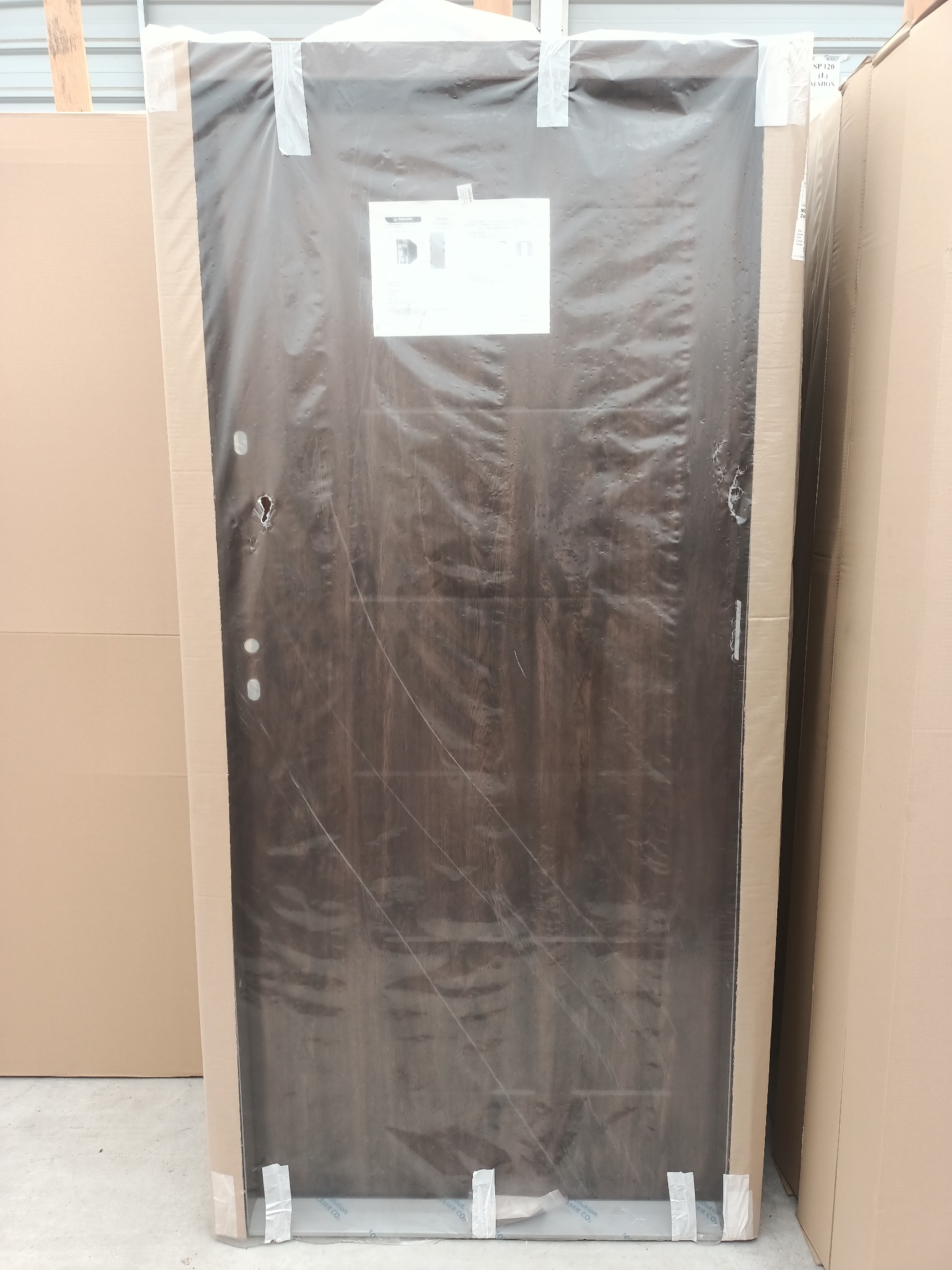 Drzwi zewnętrzne w opakowaniu ochronnym z folii, zabezpieczone taśmą klejącą, widoczny fragment etykiety informacyjnej