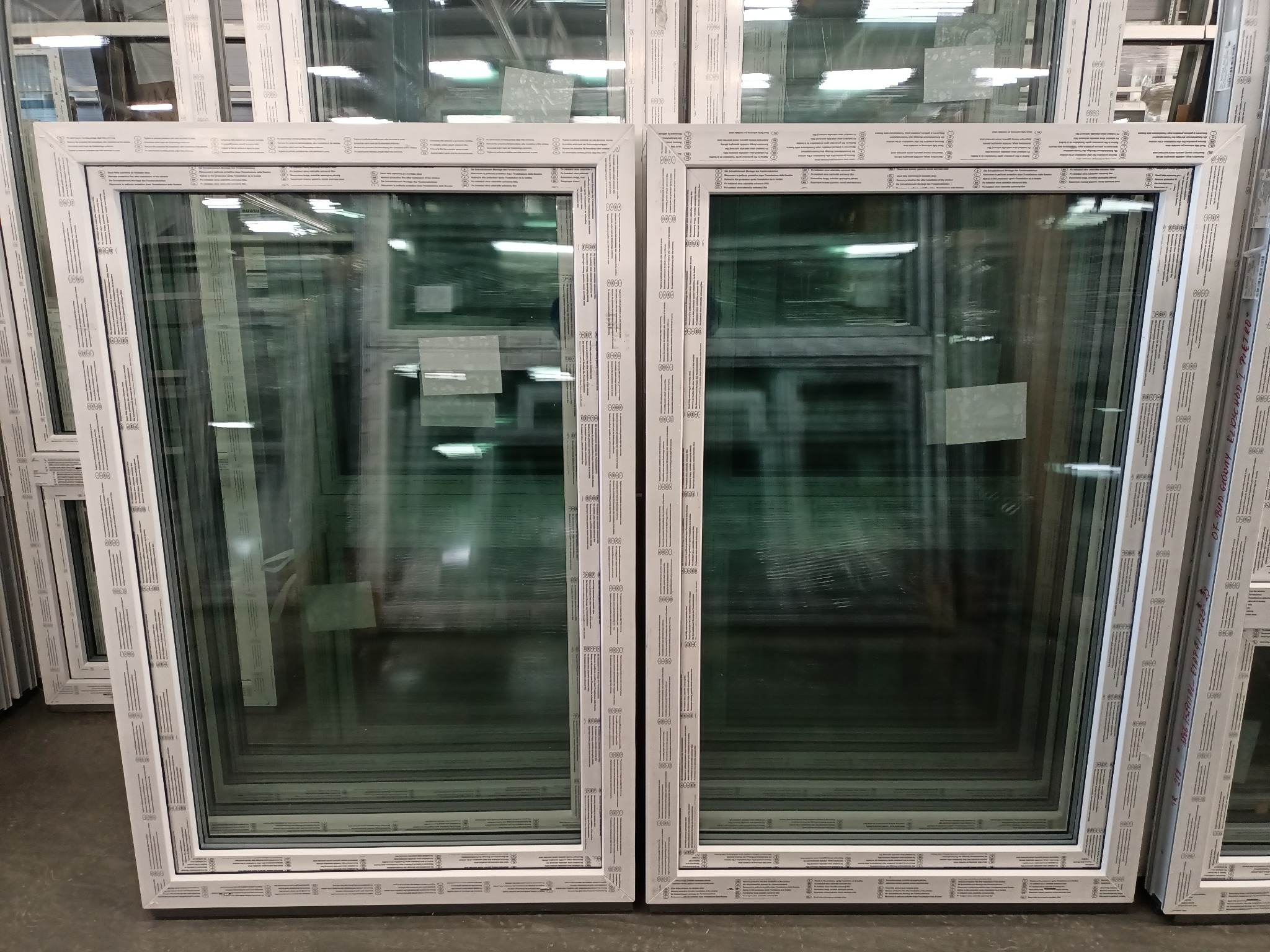 Białe ramy okienne z PCV, ułożone w stos, widoczne szyby z odblaskami magazynowego oświetlenia.