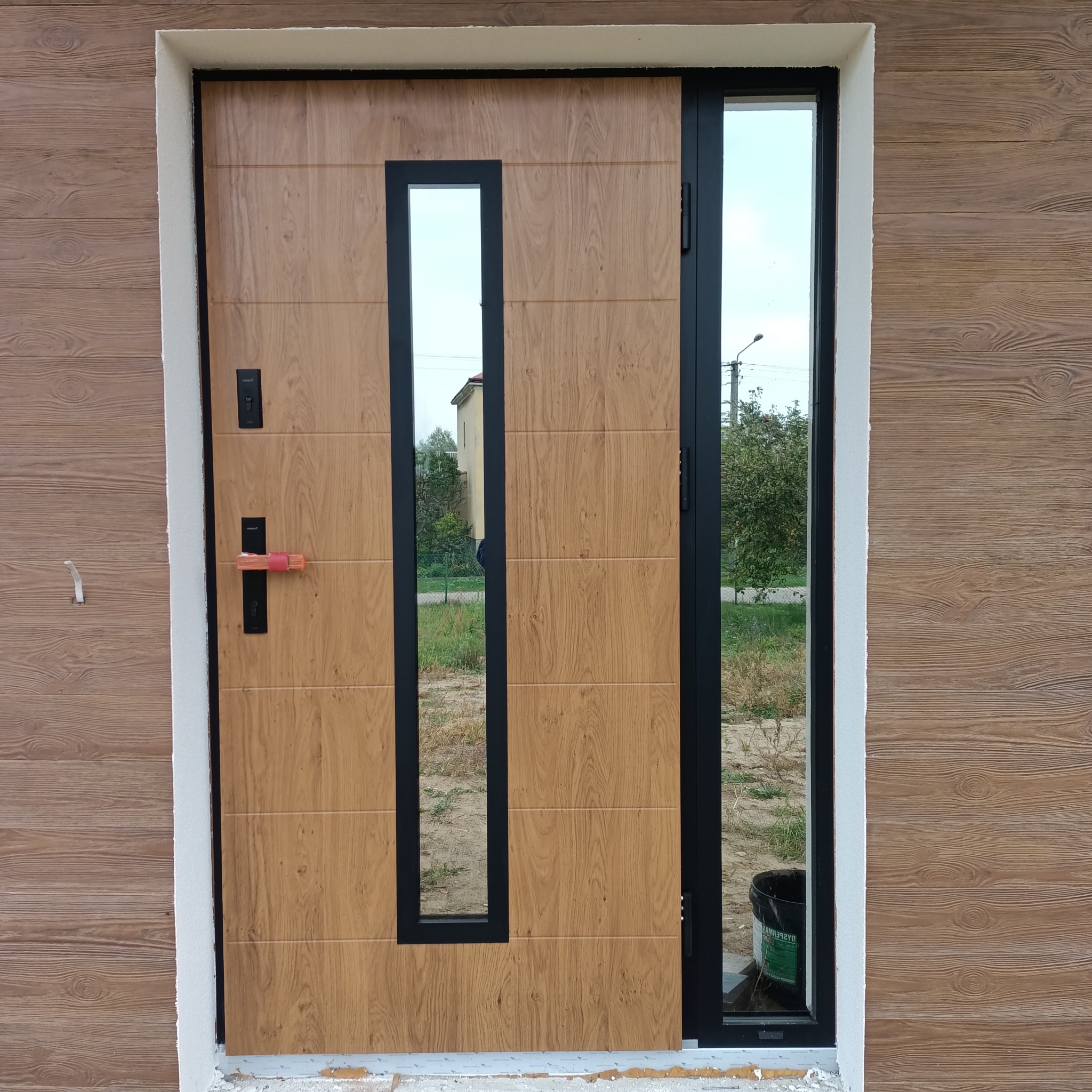 Zewnętrzne drzwi wejściowe z drewnopodobnym panelem i czarną ramą, z bocznym przeszkleniem, zamontowane w elewacji z drewnopodobnych paneli, widoczny fragment ogrodu w odbiciu szyby.