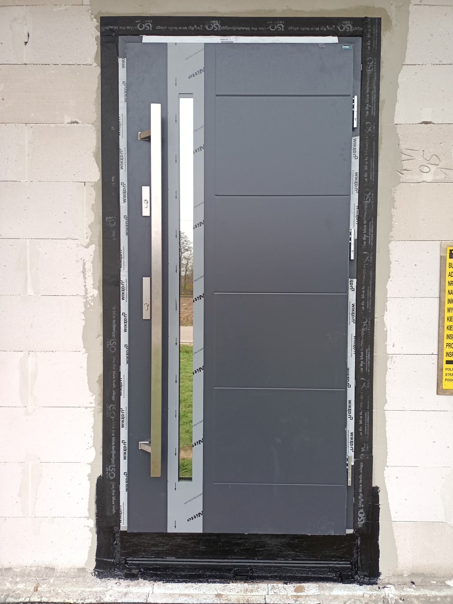 Nowoczesne, szare drzwi zewnętrzne z długim, srebrnym uchwytem, zamontowane w surowej ścianie z betonu komórkowego, zabezpieczone taśmą uszczelniającą.