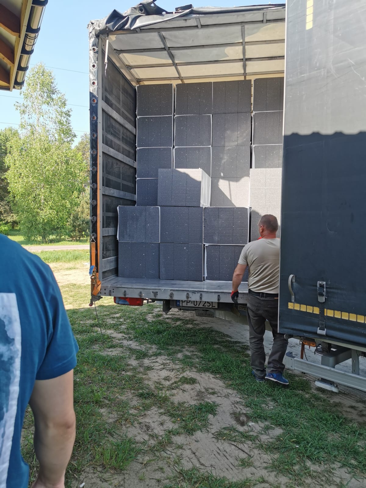 Rozładunek szarych bloków ociepleniowych z naczepy ciężarówki przez pracownika w rękawicach roboczych, widoczne tablice rejestracyjne pojazdu.