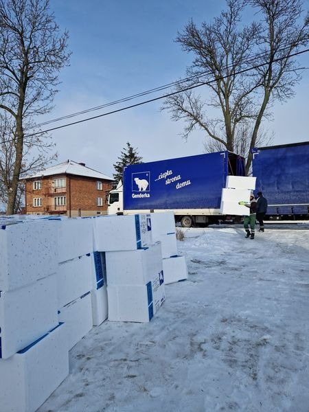 Białe bloki materiału izolacyjnego ułożone na śniegu przed ciężarówką z napisem 'ciepła strona domu' i logiem niedźwiedzia polarnego, w tle budynek mieszkalny i drzewa.
