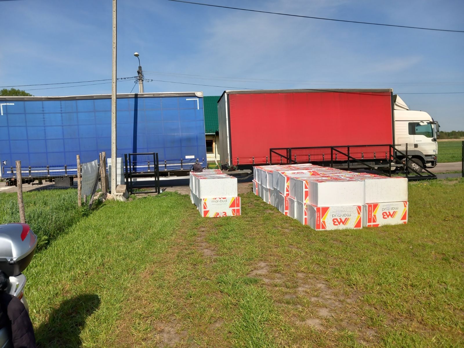 Dwa samochody ciężarowe z naczepami w kolorze niebieskim i czerwonym zaparkowane obok siebie. Na trawie ułożone palety z białymi paczkami materiału budowlanego z czerwono-żółtymi oznaczeniami.