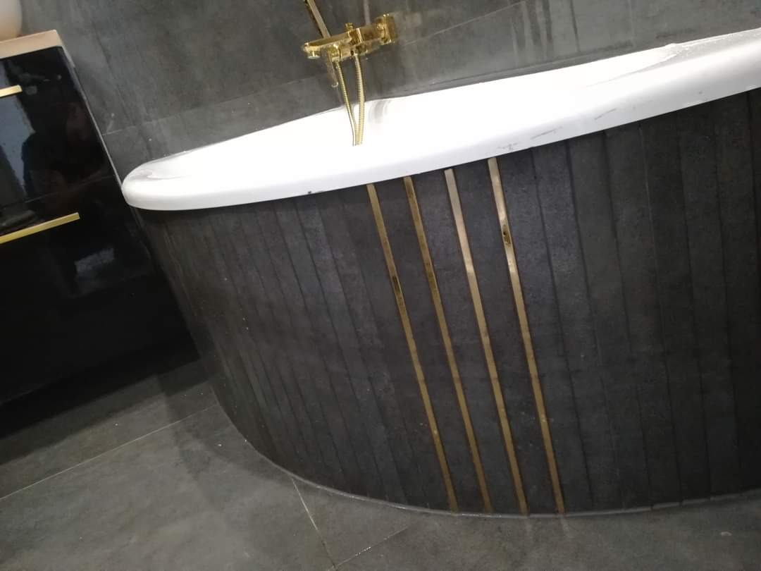 Nowoczesna wanna z obudową z ciemnych, pionowych paneli z akcentami w kolorze złotym oraz baterią w tym samym kolorze, ustawiona w łazience wykończonej szarą płytką.