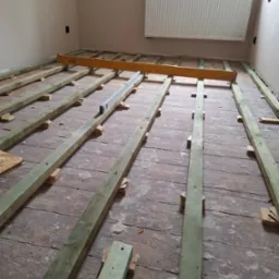 Przygotowanie podłoża pod nową podłogę: drewniane legary ułożone na starej, nierównej podłodze, poziomowane przy użyciu poziomicy i długiej łaty, widoczne ślady po starym parkiecie.