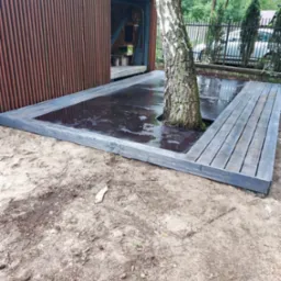 Szara drewniana platforma zbudowana wokół pnia brzozy na piaszczystym podłożu, obok rustykalnej, brązowej, metalowej konstrukcji z otwartymi drzwiami i widocznym warsztatem w tle.