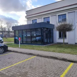 Nowoczesna, przeszklona dobudówka do budynku biurowego z widocznym parkingiem i drzewem ozdobnym.