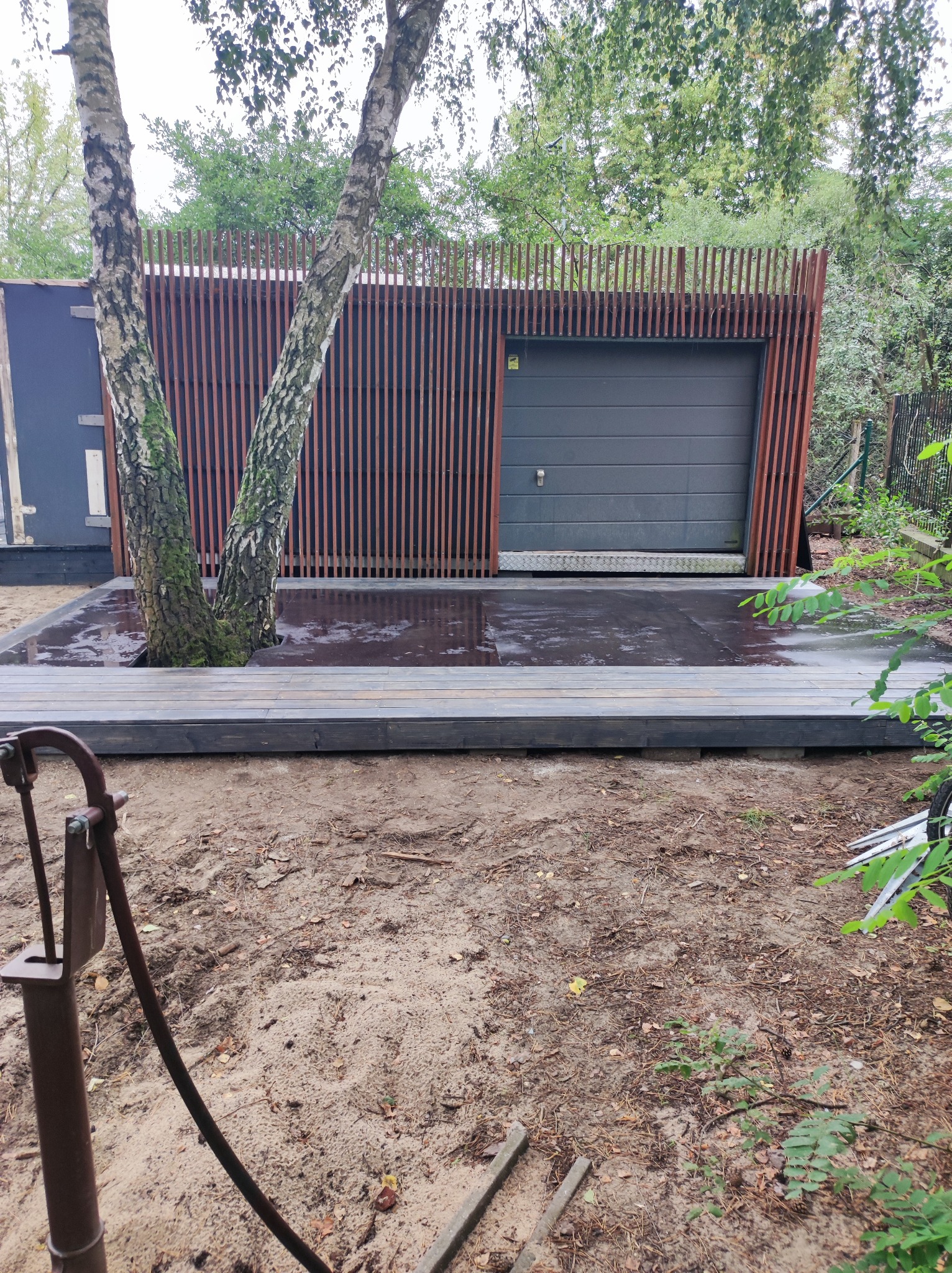 Nowoczesna altana z szarej bramy garażowej i pionowych drewnianych listew, z drzewem rosnącym wewnątrz platformy z desek, widok na piaszczyste podłoże i starą pompę wodną.