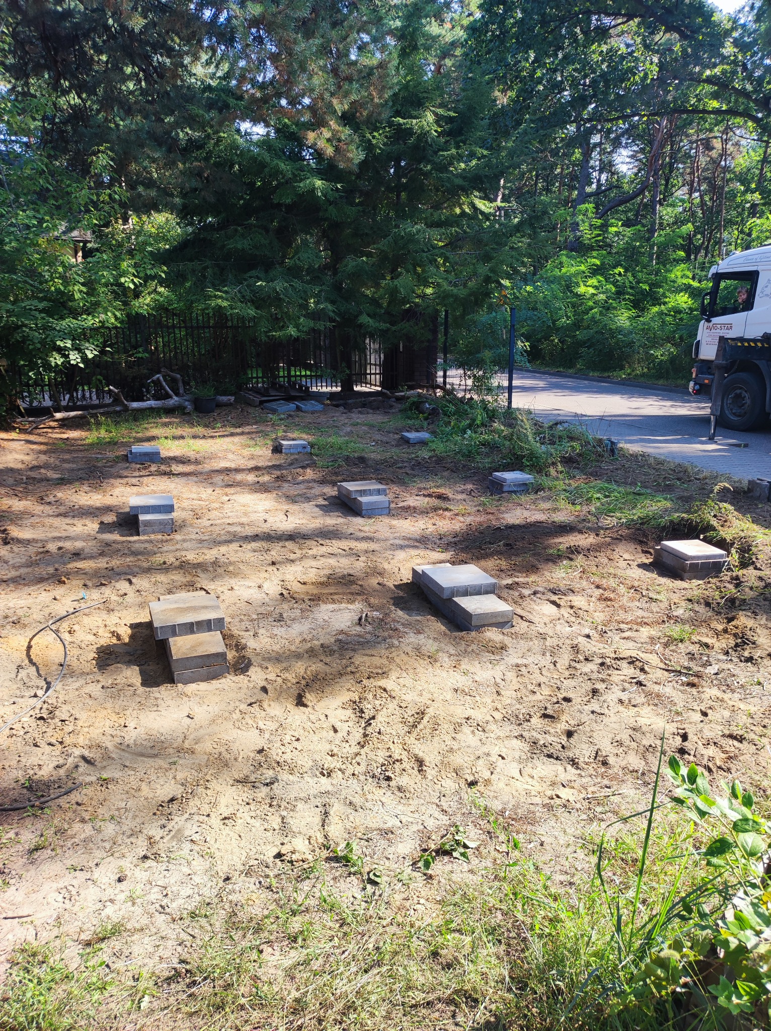 Przygotowanie podłoża pod taras kompozytowy: ułożone betonowe bloczki w wykopanym gruncie, widoczny kabel, drzewa w tle, częściowo widoczna ciężarówka.