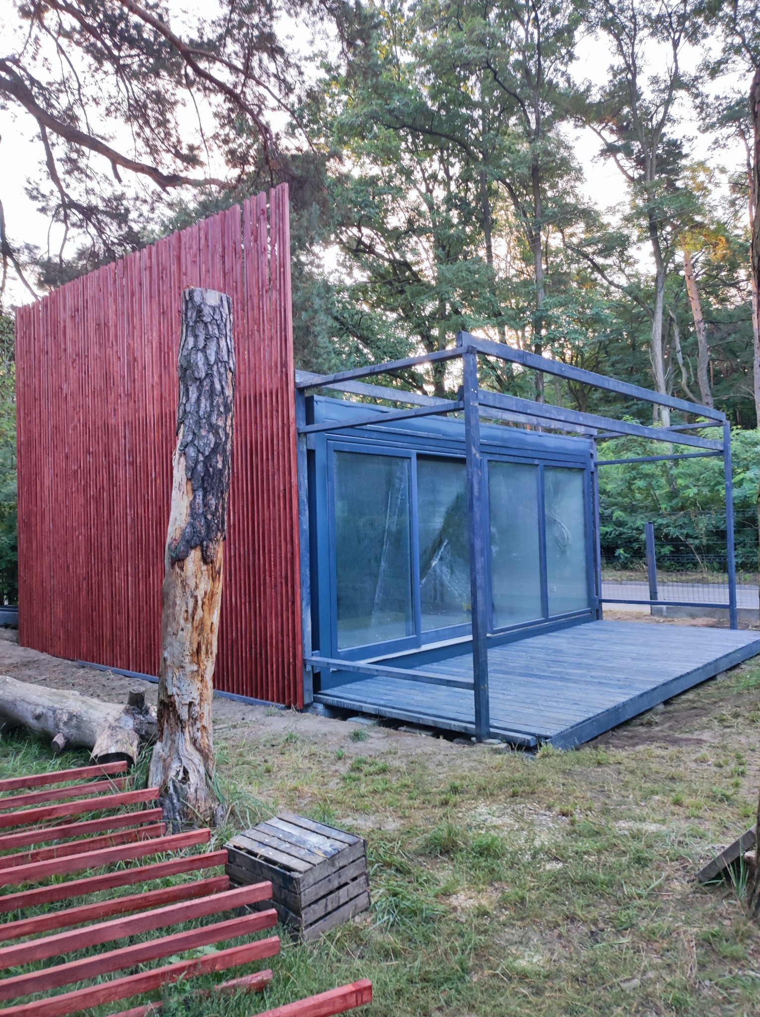 Nowoczesna altana z przeszklonymi ścianami i drewnianym tarasem, otoczona drzewami, z surowym pniem drzewa w pobliżu czerwonej, pionowej drewnianej ściany.