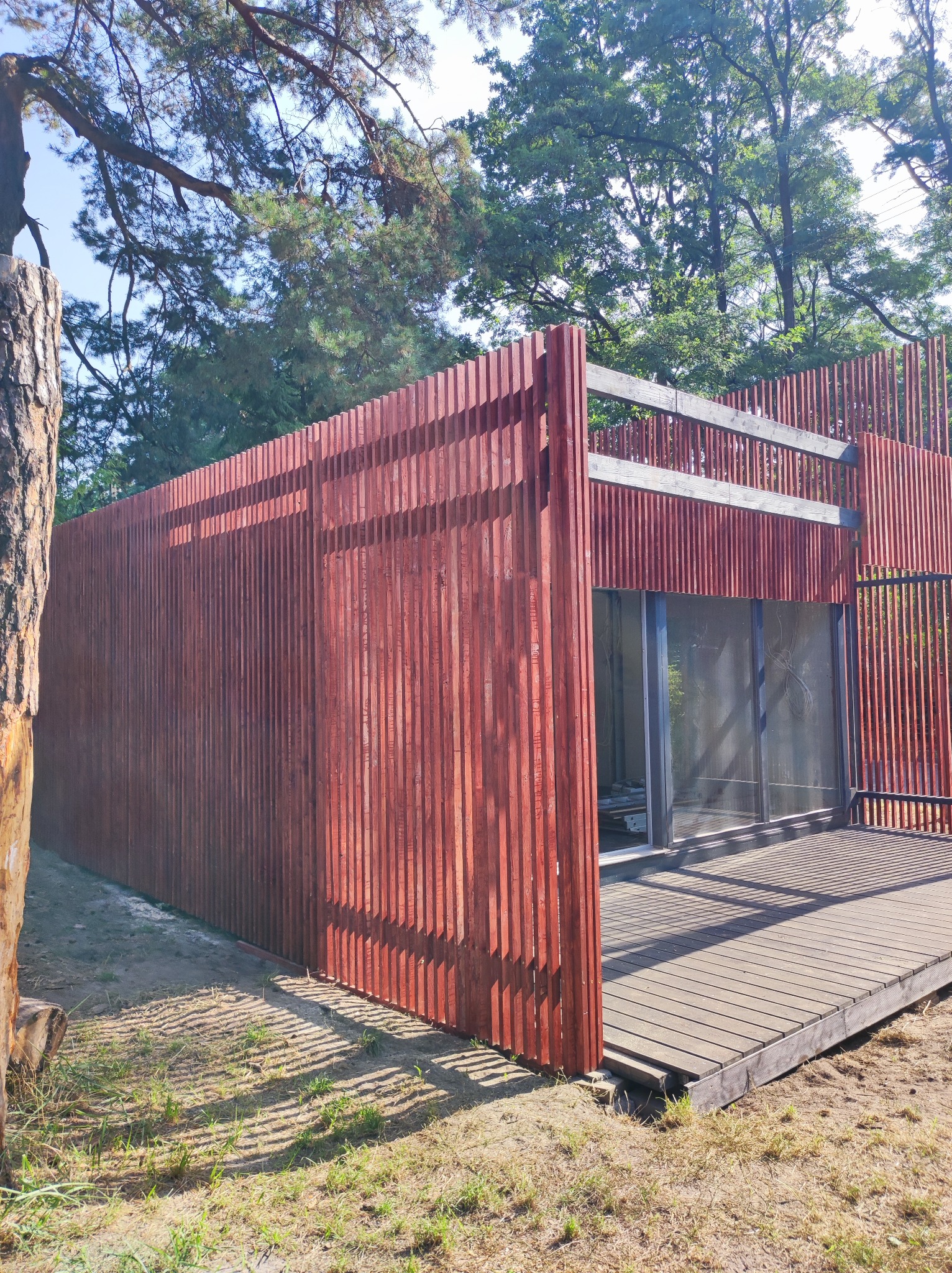 Nowoczesna konstrukcja z pionowych, lakierowanych na czerwono desek, tworząca ścianę i częściowo zadaszenie nad drewnianym tarasem, otoczona zielenią i drzewami.