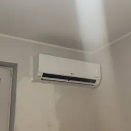 Biały klimatyzator LG zamontowany na ścianie w pokoju, widoczny z boku okna z widokiem na miasto w pochmurny dzień.