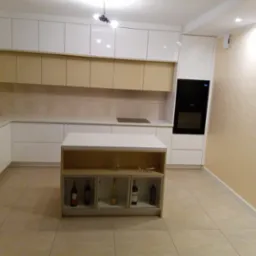 Nowoczesna kuchnia z białymi i beżowymi szafkami, wyspą z ekspozycją na butelki wina, piekarnikiem w zabudowie i beżowymi płytkami na ścianie.