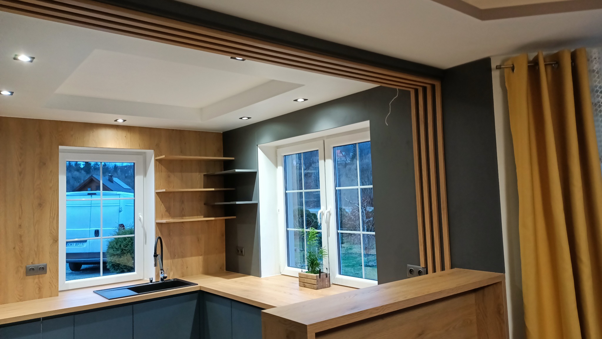 Nowoczesna kuchnia z drewnianymi akcentami: blat zlewem i otwartymi półkami, okno z widokiem na zewnątrz, dekoracyjne listwy na ścianie, żółte zasłony.