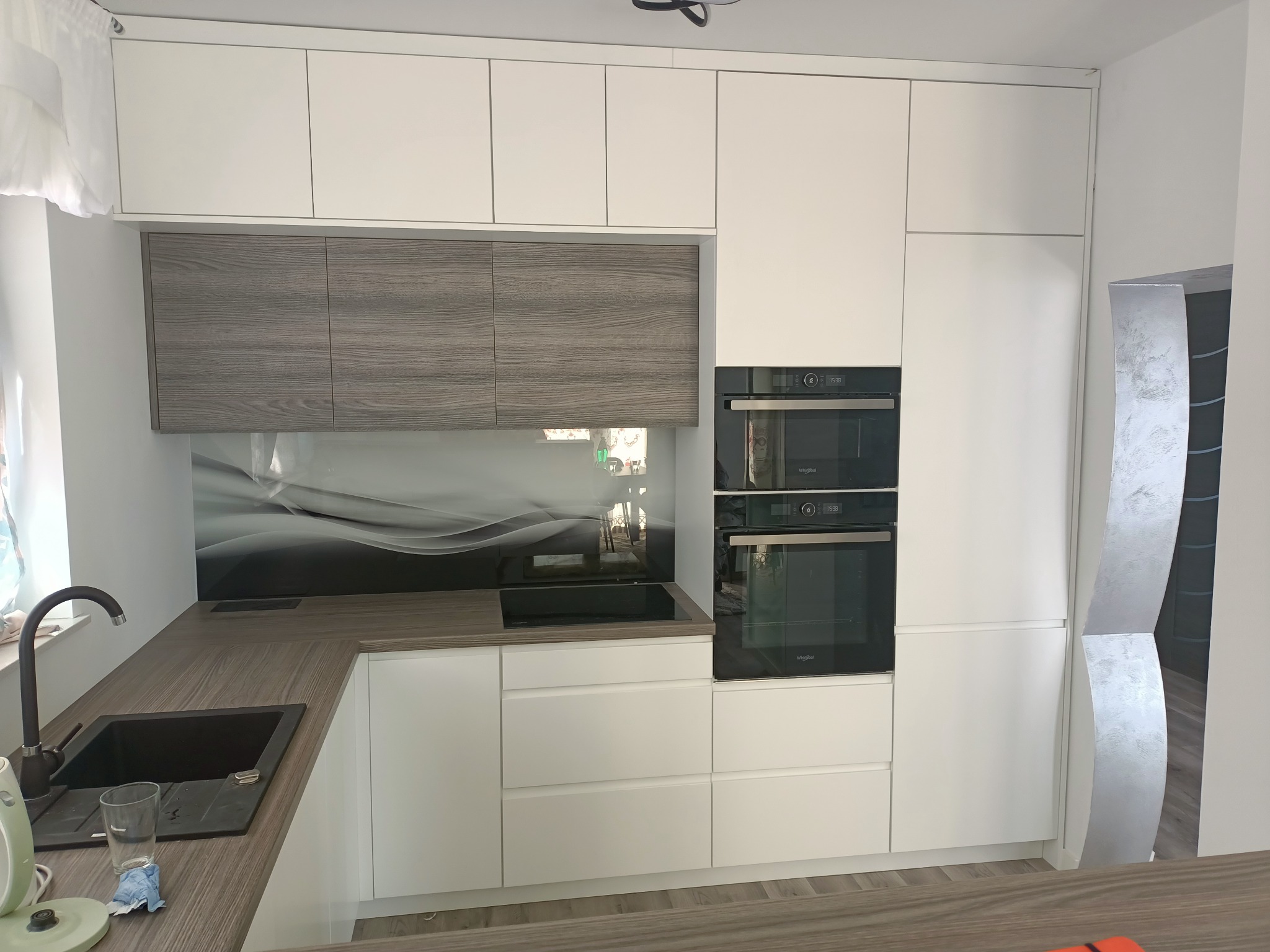 Nowoczesna kuchnia z białymi frontami szafek i blatem w kolorze drewna, czarny zlew i płyta indukcyjna, szklany panel nad blatem z motywem fal, zabudowane piekarniki.