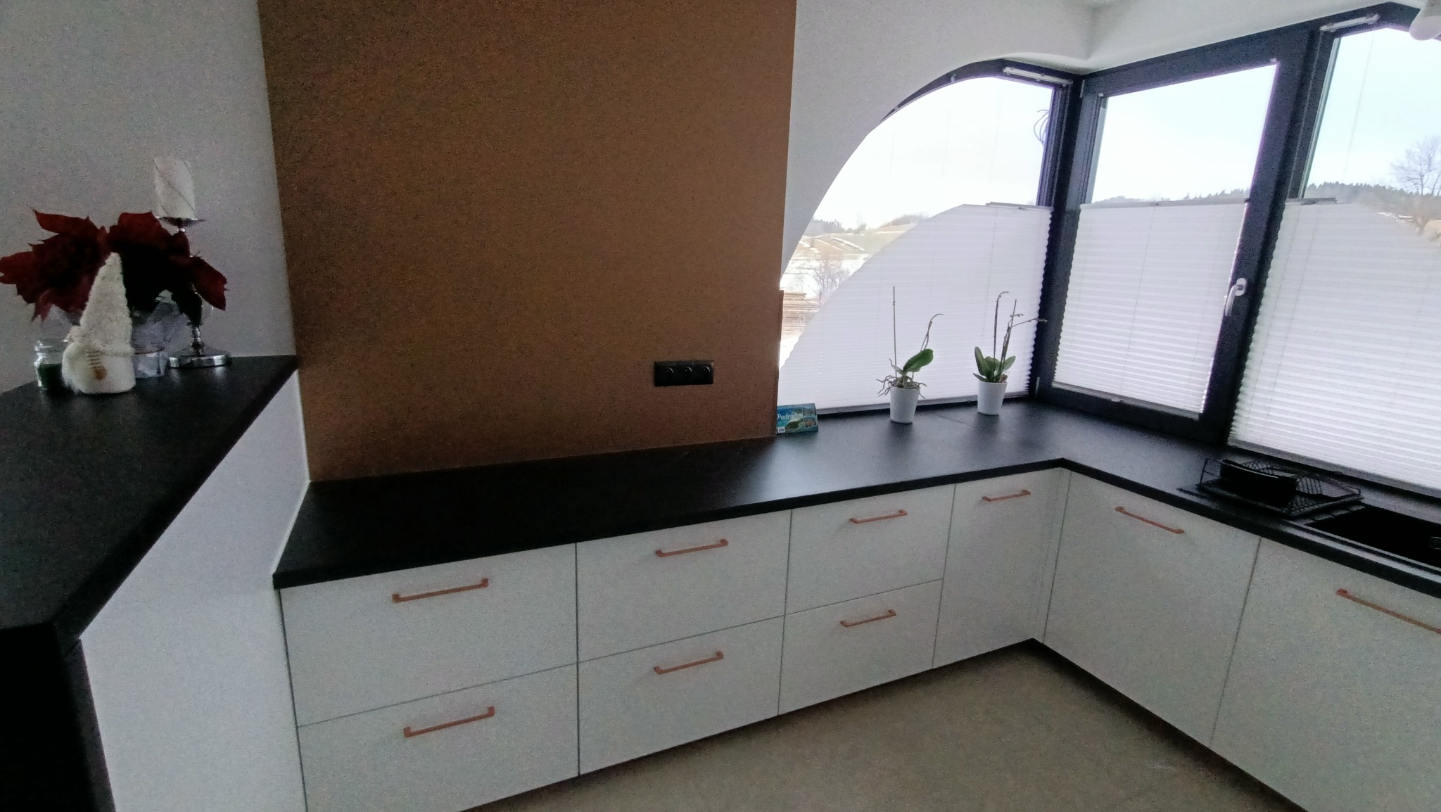 Białe szafki kuchenne z miedzianymi uchwytami pod ciemnym blatem, dekoracje i rośliny na blacie, okno z plisowanymi roletami w tle.