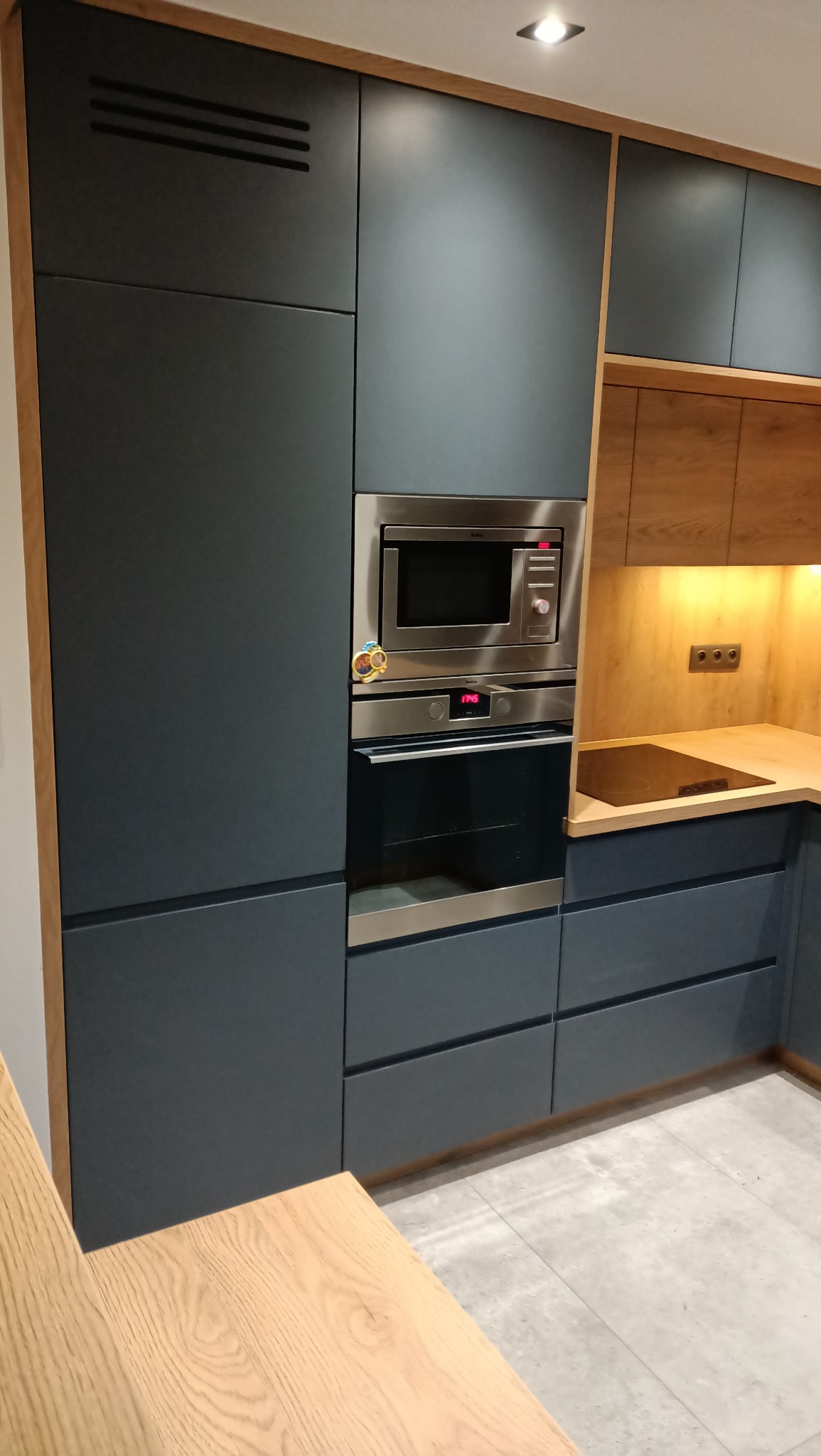 Nowoczesna zabudowa kuchenna w odcieniach szarości z drewnianymi akcentami, obejmująca wbudowany piekarnik i mikrofalówkę, oświetlona punktowo.