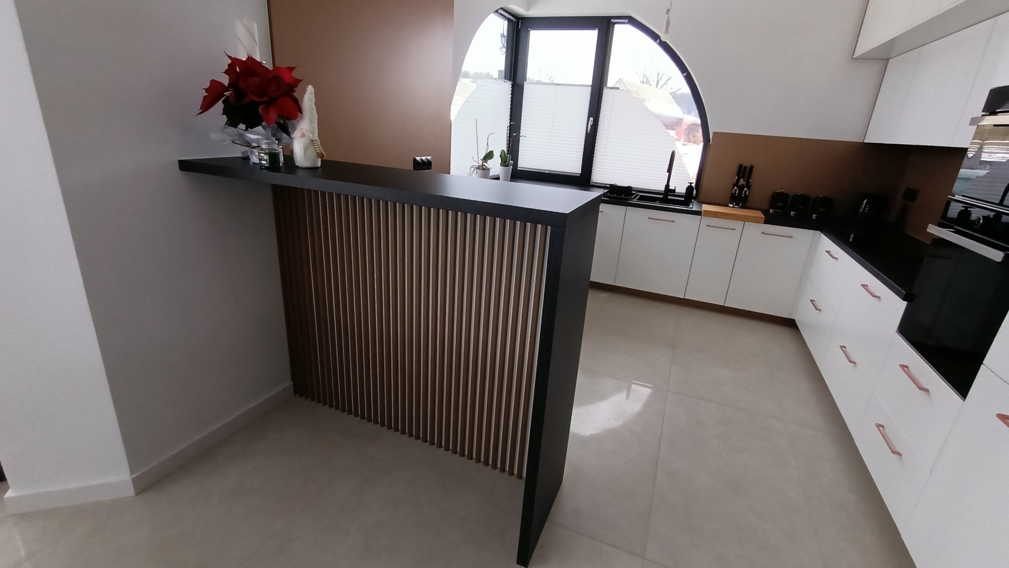 Nowoczesna kuchnia z białymi szafkami, czarnym blatem i barkiem z drewnianymi lamelami, w tle okno z roletami plisowanymi.