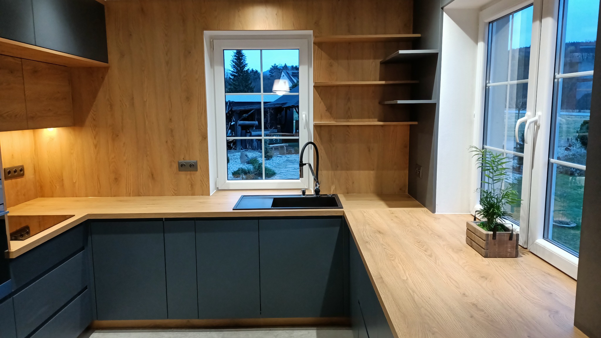 Nowoczesna kuchnia z drewnianymi blatami i frontami w kolorze ciemnego grafitu, czarnym zlewem pod oknem z widokiem na ogród, oraz drewnianymi półkami na ścianie.