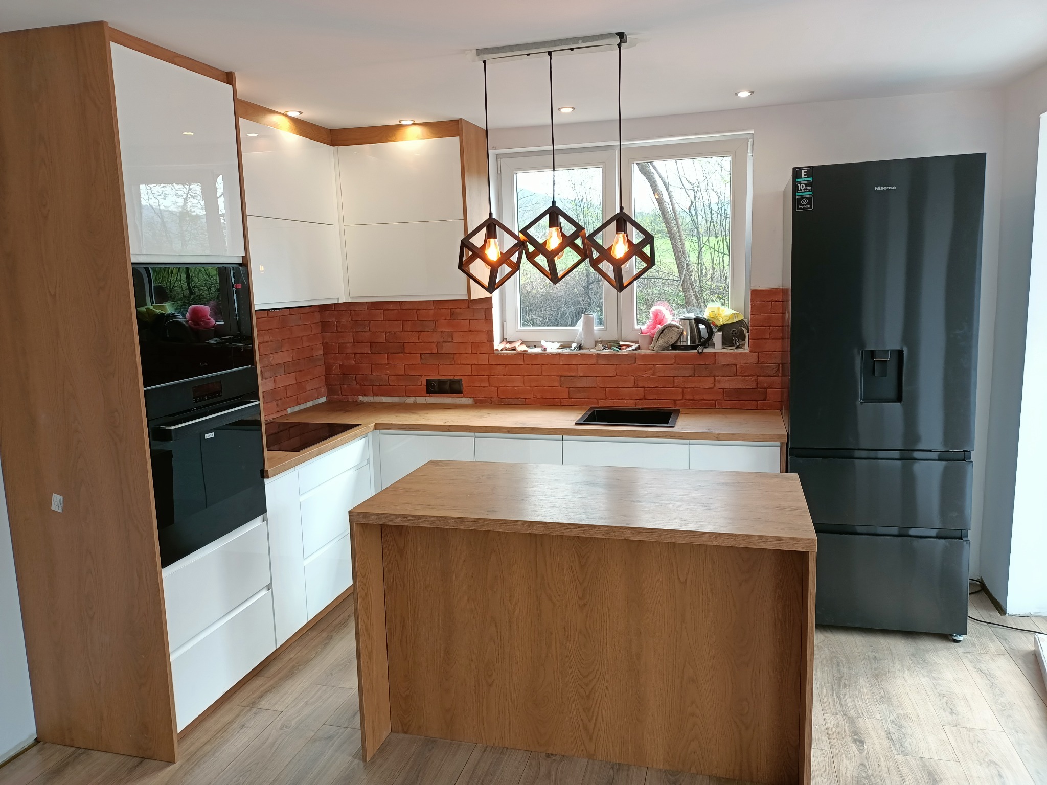 Nowoczesna kuchnia z białymi szafkami, drewnianymi blatami i wyspą, ceglaną ścianą nad blatem oraz geometrycznymi lampami wiszącymi w kształcie sześcianów, uzupełniona czarną lodówką.