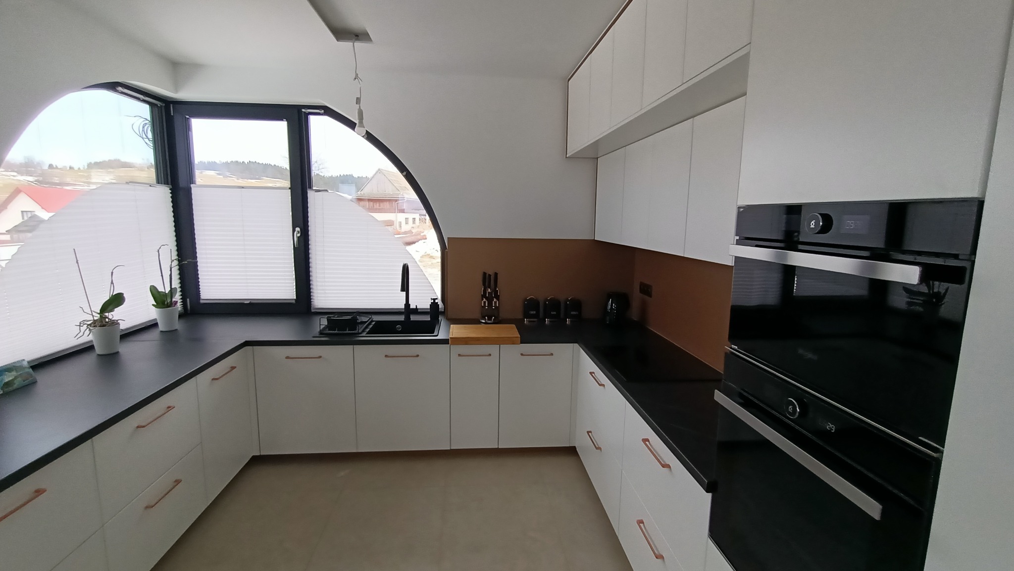 Nowoczesna kuchnia z białymi szafkami, czarnym blatem i miedzianymi uchwytami, nietypowe okno z roletami plisowanymi, zabudowany piekarnik i brązowy panel nad blatem.