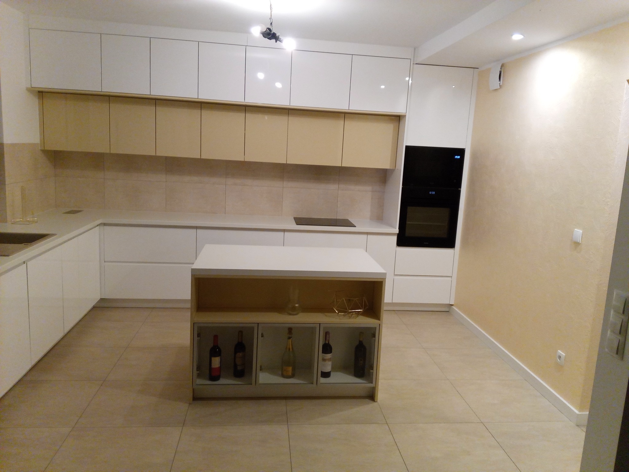 Nowoczesna kuchnia z białymi i beżowymi szafkami, wyspą z ekspozycją na butelki wina, piekarnikiem w zabudowie i beżowymi płytkami na ścianie.