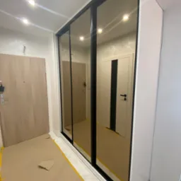 Wnękowa szafa z lustrzanymi drzwiami przesuwnymi i czarnymi ramami, widoczna z zabezpieczonym podłożem w trakcie montażu.