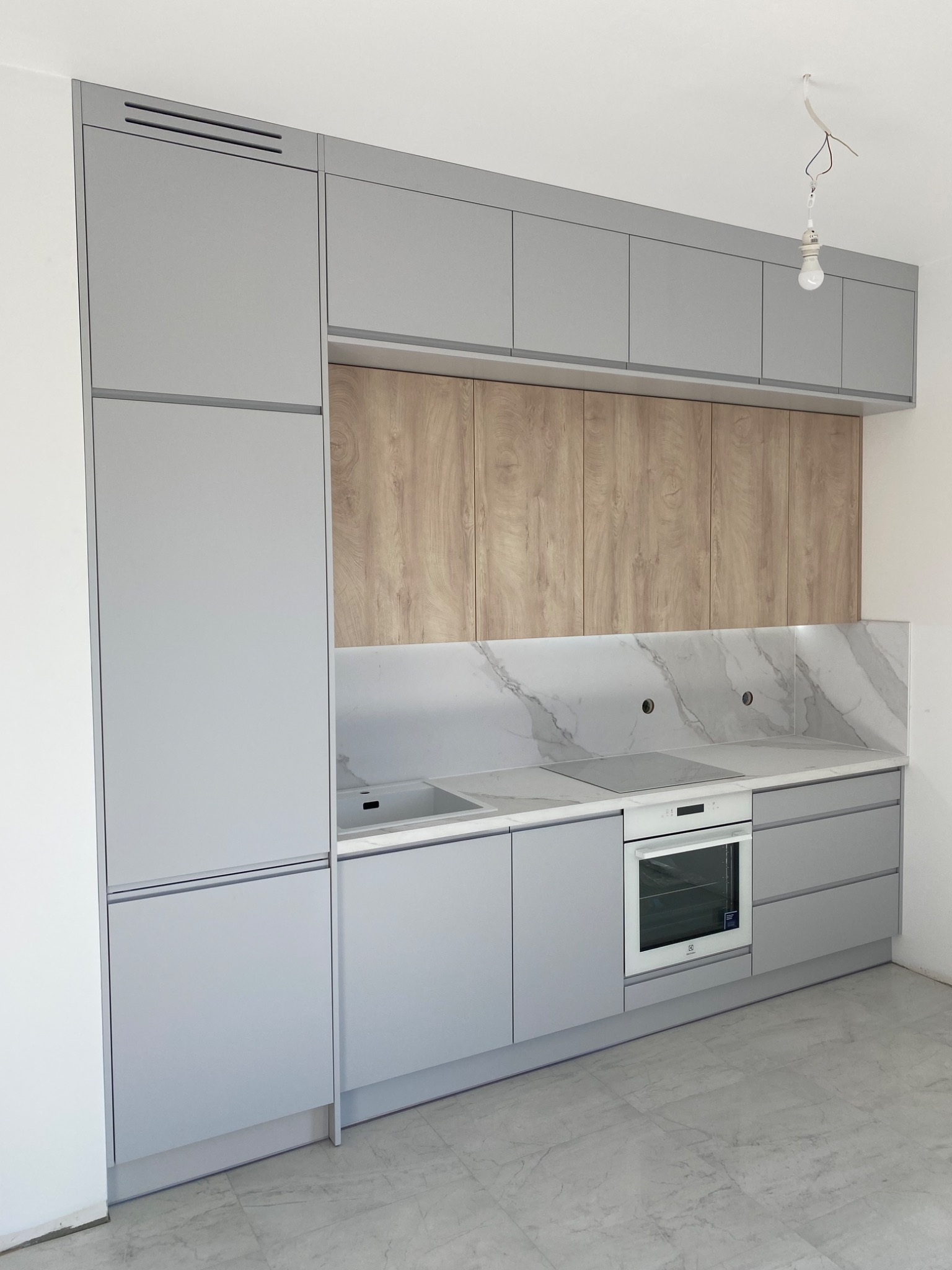 Szara zabudowa kuchenna z drewnianymi szafkami górnymi i marmurowym blatem, widoczna niedokończona instalacja oświetleniowa.