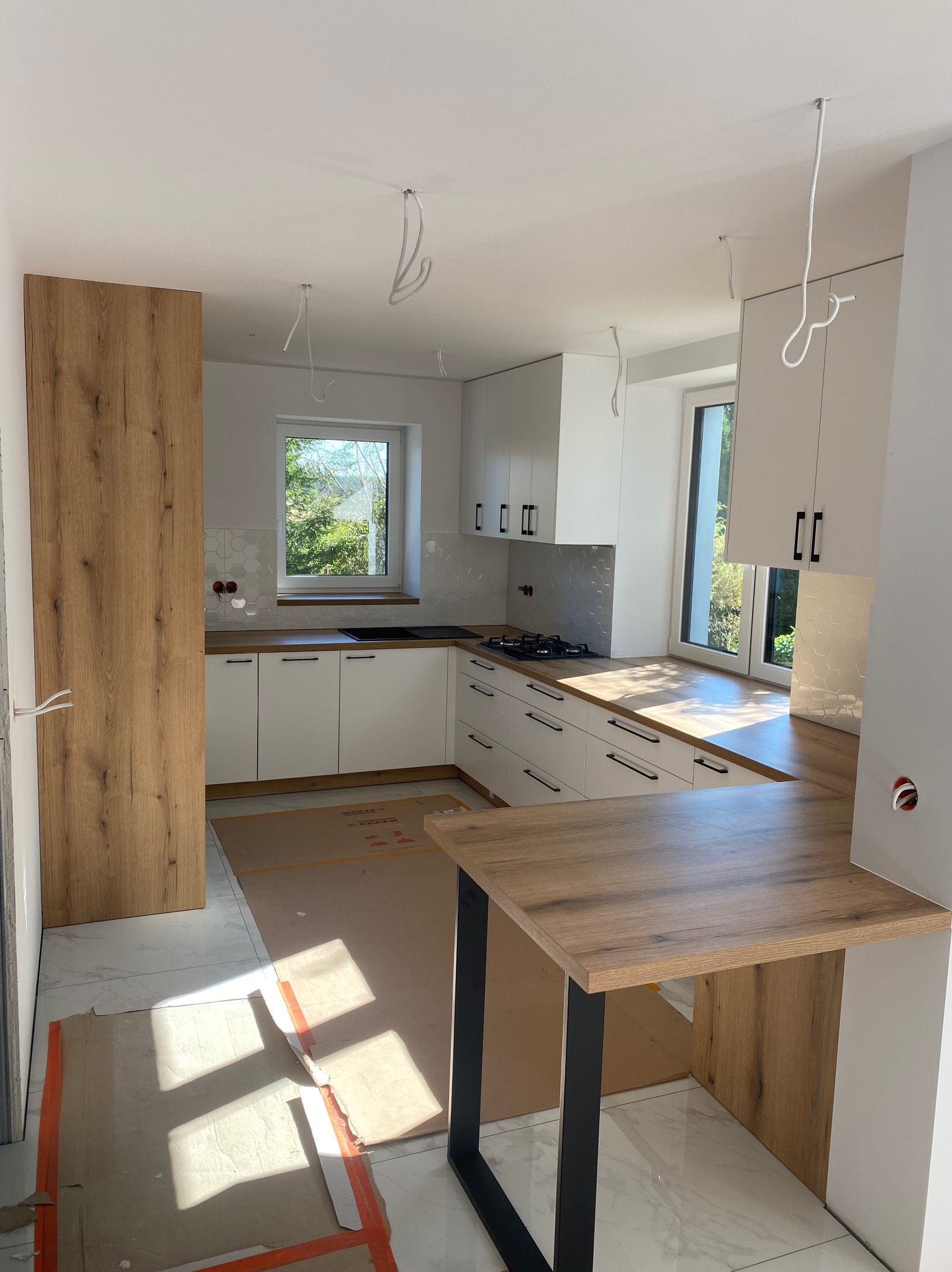 Jasna kuchnia w trakcie montażu: białe szafki z czarnymi uchwytami, drewniany blat i wyspa, na podłodze zabezpieczenia kartonowe, wystające kable oświetleniowe.