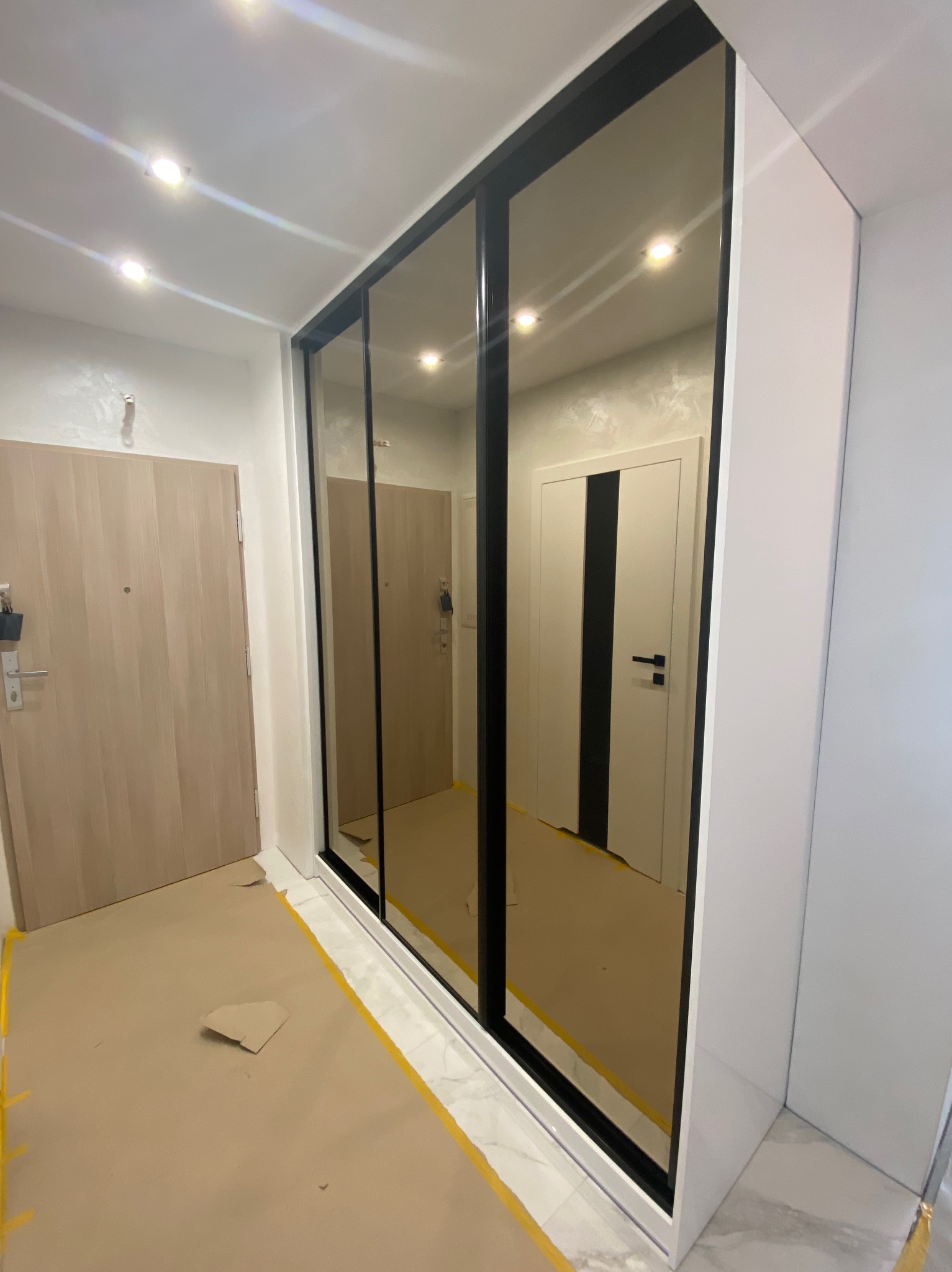 Wnękowa szafa z lustrzanymi drzwiami przesuwnymi i czarnymi ramami, widoczna z zabezpieczonym podłożem w trakcie montażu.