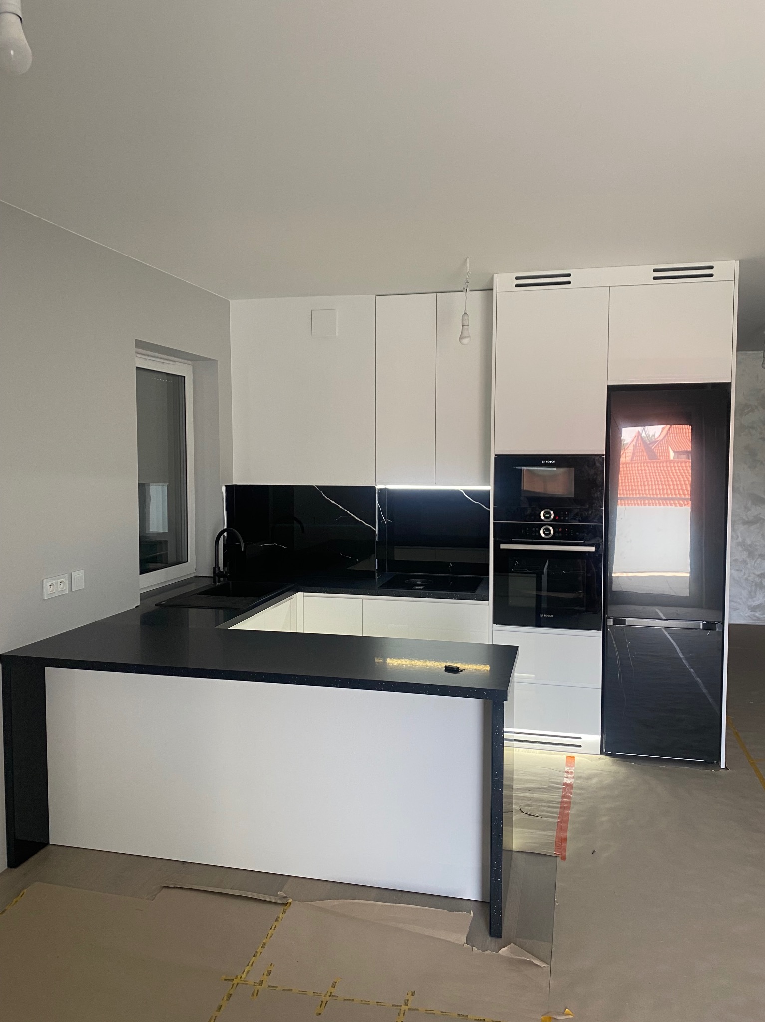 Nowoczesna kuchnia z białymi szafkami i czarnym blatem z marmurowym wzorem, wyspa kuchenna, wbudowany piekarnik i lodówka, widoczne zabezpieczenia podłogi podczas montażu.