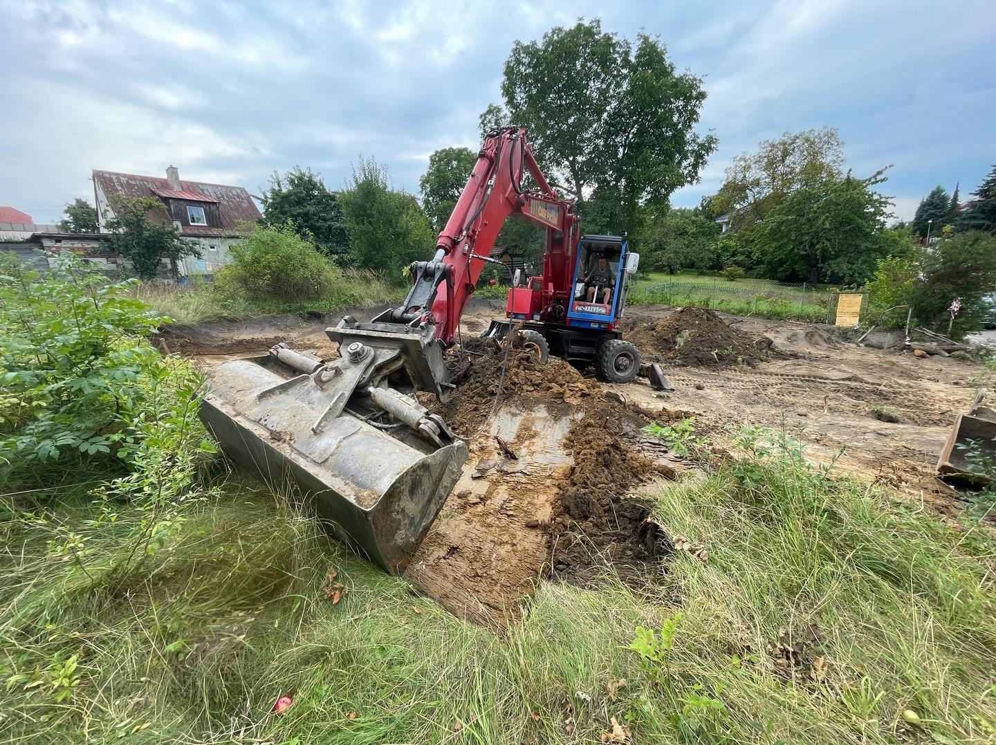 Czerwono-niebieska koparka kołowa z łyżką zagłębia się w ziemię na placu budowy, w tle zielona roślinność i budynek mieszkalny pod pochmurnym niebem.