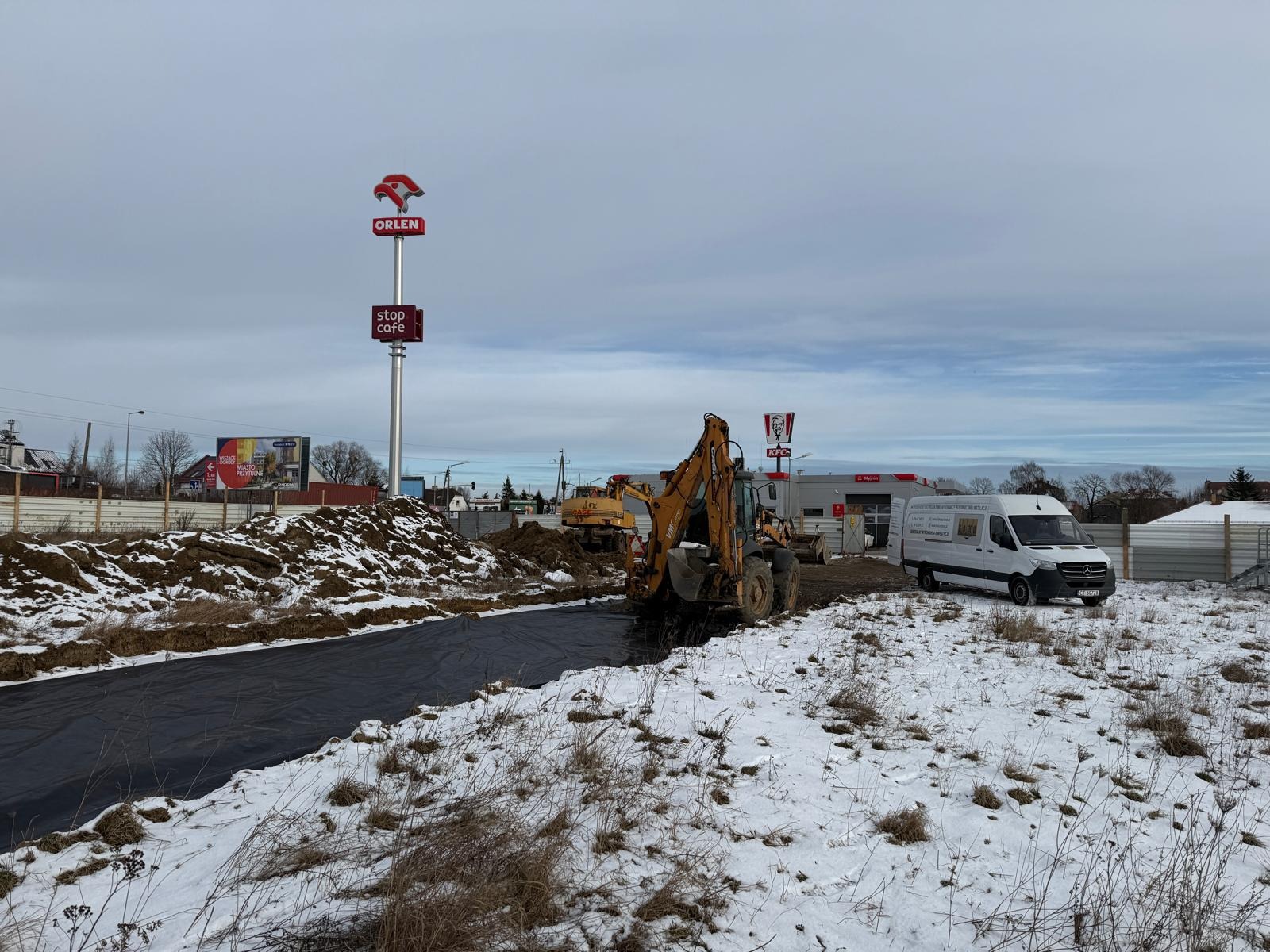 Prace ziemne w zimowej scenerii: koparka Case i spycharka w tle, czarna folia na wykopie. W tle stacja Orlen i KFC.