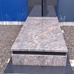 B.U. REM-BUD BOGUSŁAW BUCZKO - Granitowy pomnik z dwoma pionowymi płytami, ustawiony na żwirowym podłożu przed niebieską ścianą z blachy falistej.