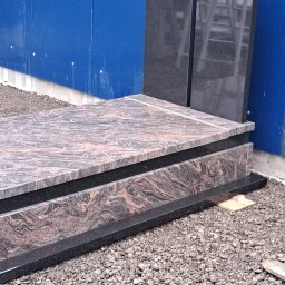 B.U. REM-BUD BOGUSŁAW BUCZKO - Granitowy pomnik z ciemnoróżowymi żyłkami, ustawiony na żwirowym podłożu przed niebieską ścianą z blachy falistej.