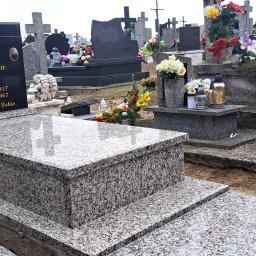 B.U. REM-BUD BOGUSŁAW BUCZKO - Szary granitowy nagrobek z czarną tablicą nagrobną z napisem i krucyfiksem, widok z perspektywy ziemi na cmentarzu, w tle inne nagrobki i kwiaty, obok nagrobka leżą środki czystości.