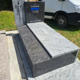 Granitowy nagrobek z krzyżem i inskrypcją 'Ś.P. Wojciech' z datami, umieszczony na cmentarzu, z białym samochodem dostawczym w tle.