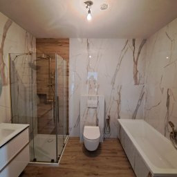 B.U. REM-BUD BOGUSŁAW BUCZKO - Wykończona łazienka z kabiną prysznicową, podwieszaną toaletą, umywalką z szafką i wanną, wyłożona płytkami imitującymi biały marmur z brązowymi żyłami oraz płytkami imitującymi drewno.