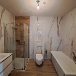 Wykończona łazienka z kabiną prysznicową, podwieszaną toaletą, umywalką z szafką i wanną, wyłożona płytkami imitującymi biały marmur z brązowymi żyłami oraz płytkami imitującymi drewno.