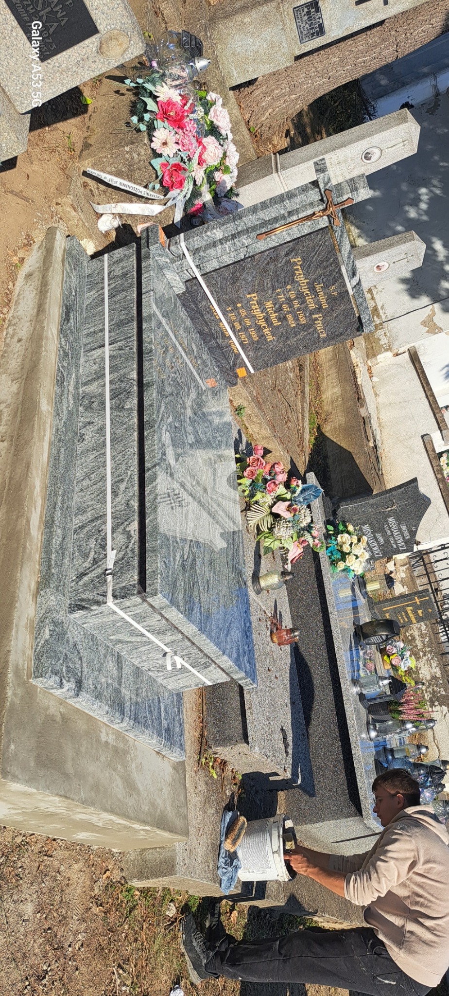 Czyszczenie jasnoszarego nagrobka z granitu o wyraźnym wzorze, widoczne kwiaty i znicze na sąsiednich grobach, osoba w trakcie pracy z wiadrem i szczotką.