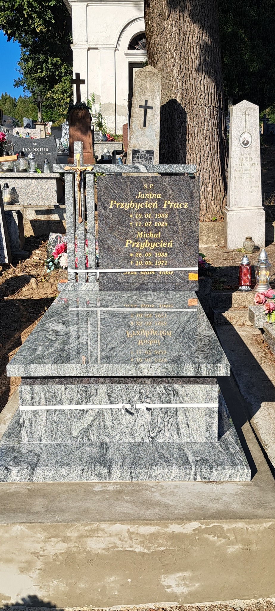 Granitowy pomnik nagrobny z inskrypcjami w złotym kolorze, otoczony innymi nagrobkami na cmentarzu, zabezpieczony białym sznurkiem.