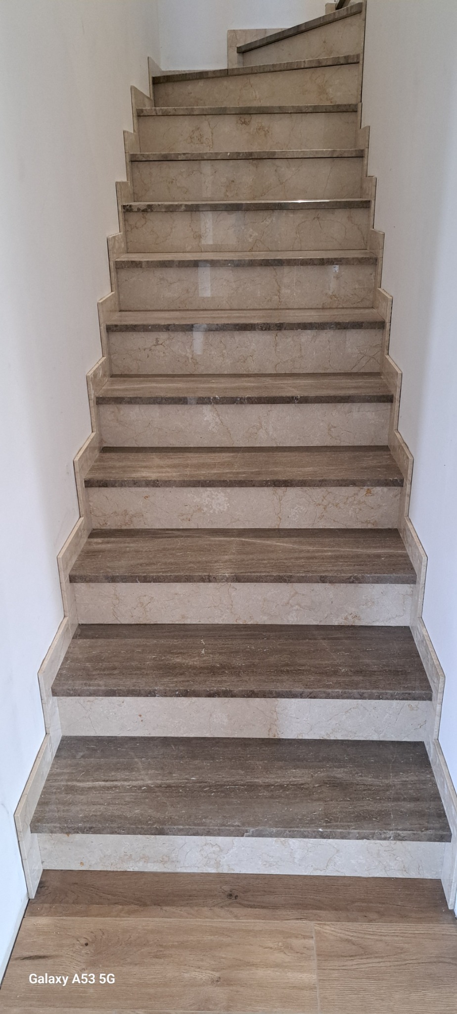 Beżowe schody z kamienia naturalnego z ciemniejszymi wstawkami, widok z dołu na górę, białe ściany po bokach.