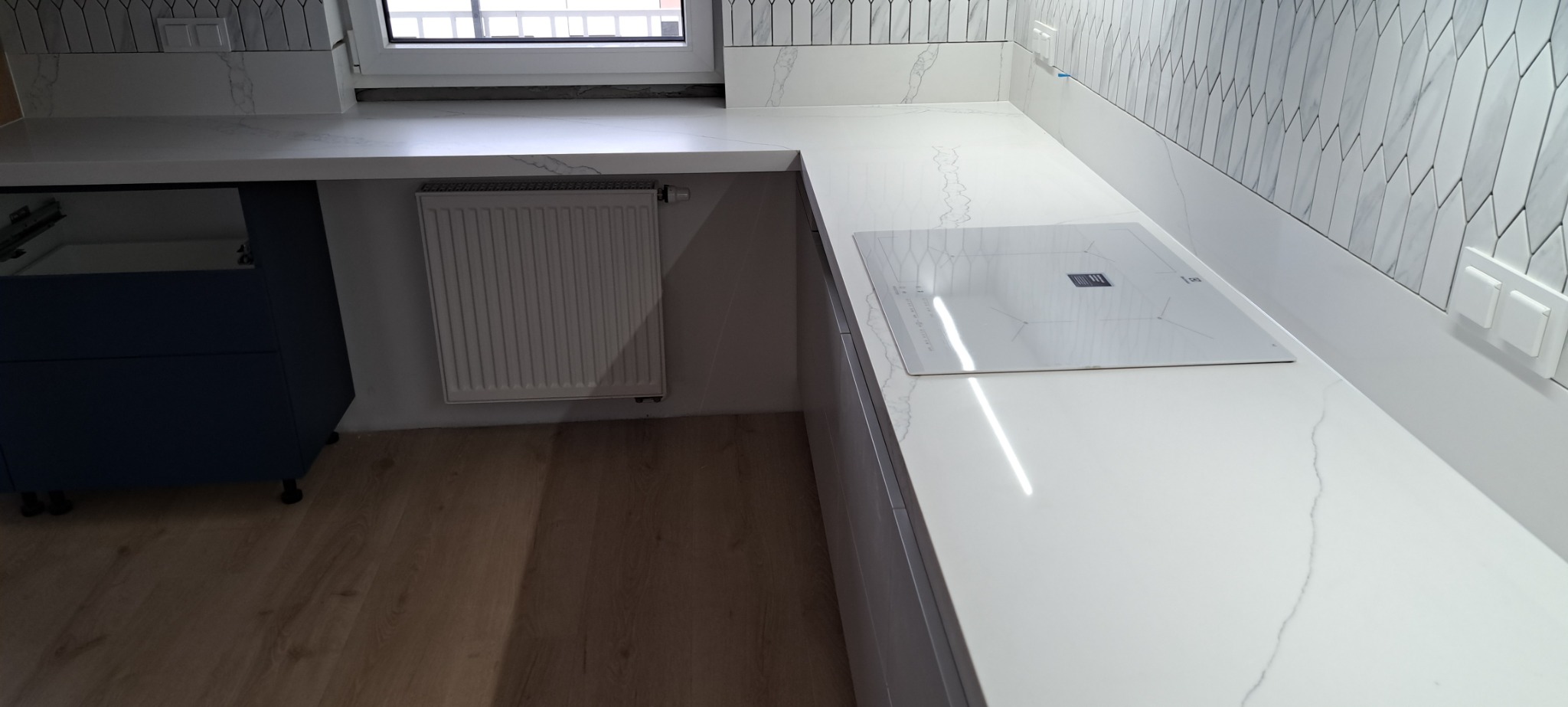 Nowoczesna kuchnia z białym blatem imitującym marmur, płyta indukcyjna i mozaika na ścianie, widok na grzejnik i fragment niebieskiej zabudowy.