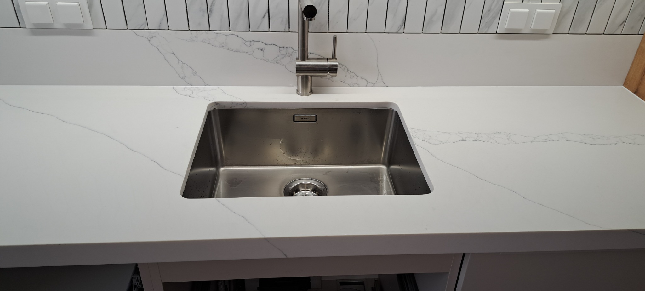 Biały blat kuchenny z konglomeratu z wbudowanym zlewozmywakiem ze stali nierdzewnej i chromowaną baterią, widoczne marmurowe płytki na ścianie.