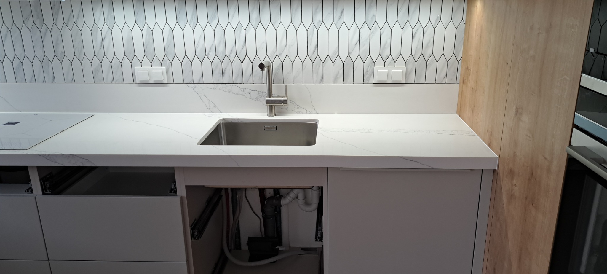 Nowoczesna kuchnia w trakcie montażu: biały blat z konglomeratu z wbudowanym zlewem, bateria, płyta indukcyjna, dekoracyjna mozaika na ścianie oraz widoczne elementy instalacji hydraulicznej...