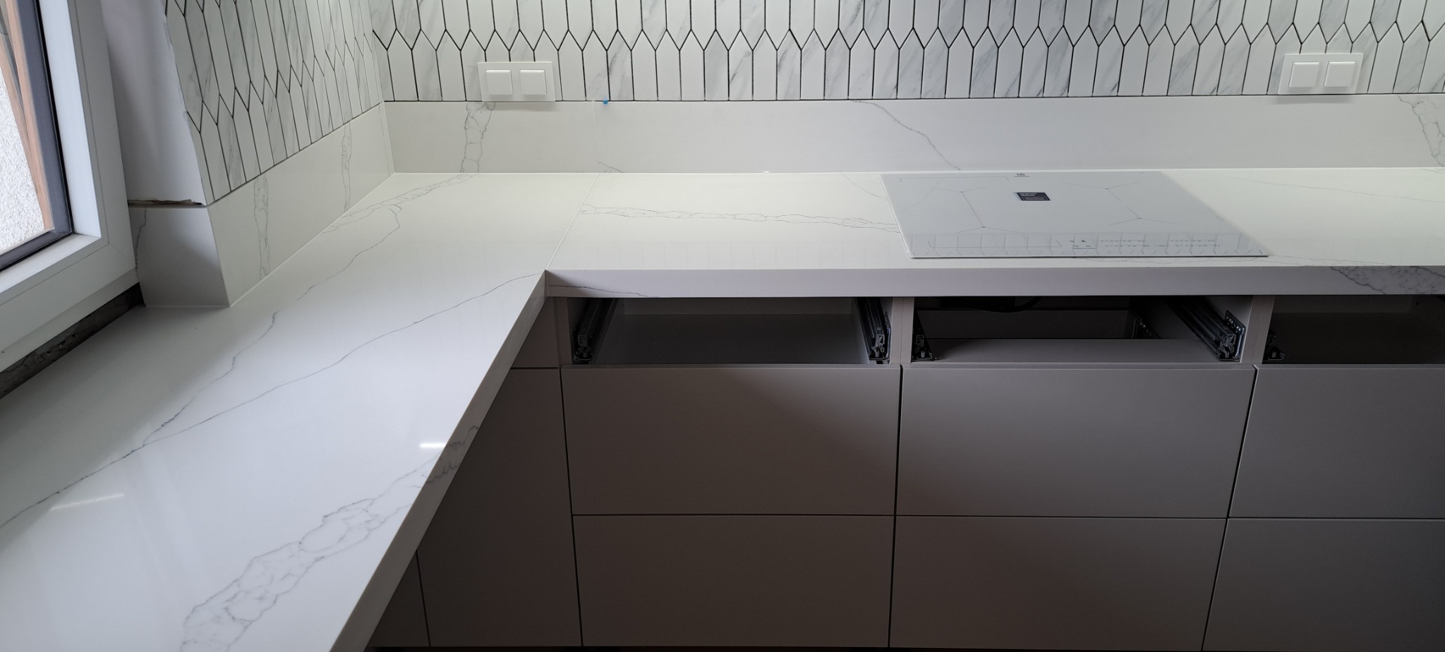 Biały blat kuchenny z konglomeratu z wbudowaną płytą indukcyjną, otwarte szuflady z widocznymi prowadnicami, mozaika na ścianie nad blatem, narożne ułożenie blatu przy oknie.