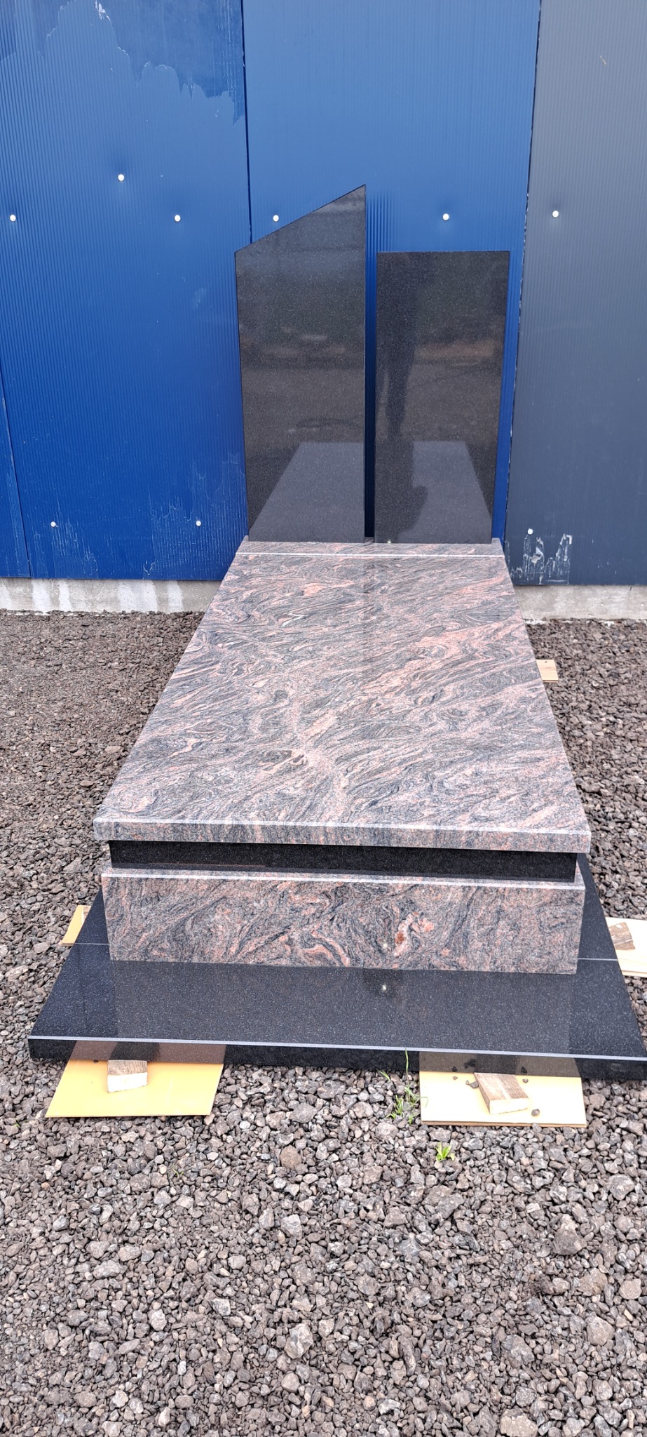Granitowy pomnik z dwoma pionowymi płytami, ustawiony na żwirowym podłożu przed niebieską ścianą z blachy falistej.