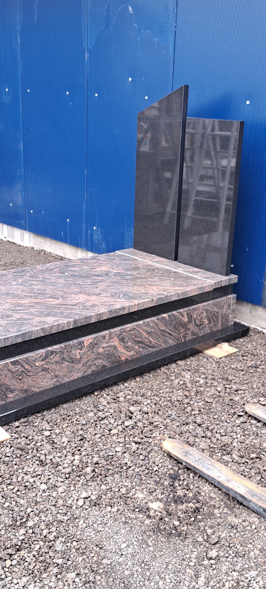 Granitowy pomnik z ciemnoróżowymi żyłkami, ustawiony na żwirowym podłożu przed niebieską ścianą z blachy falistej.