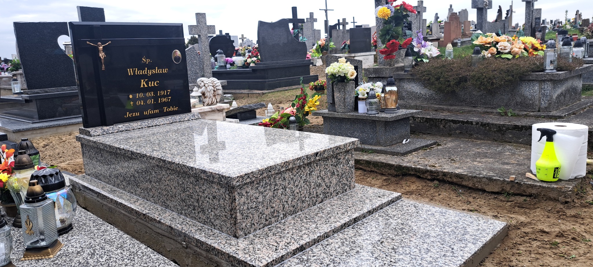 Szary granitowy nagrobek z czarną tablicą nagrobną z napisem i krucyfiksem, widok z perspektywy ziemi na cmentarzu, w tle inne nagrobki i kwiaty, obok nagrobka leżą środki czystości.